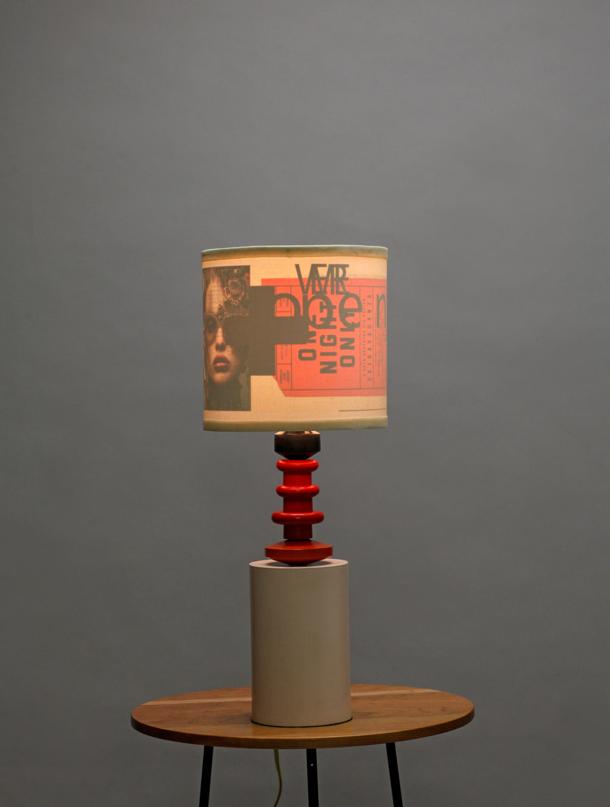 A table lamp with a beige drum shade featuring a graphic print of a face and the text 'VOTE DOCTOR NIGHT ONE', supported by a red spiral stem and a beige cylindrical base, set on a wooden table against a gray background.