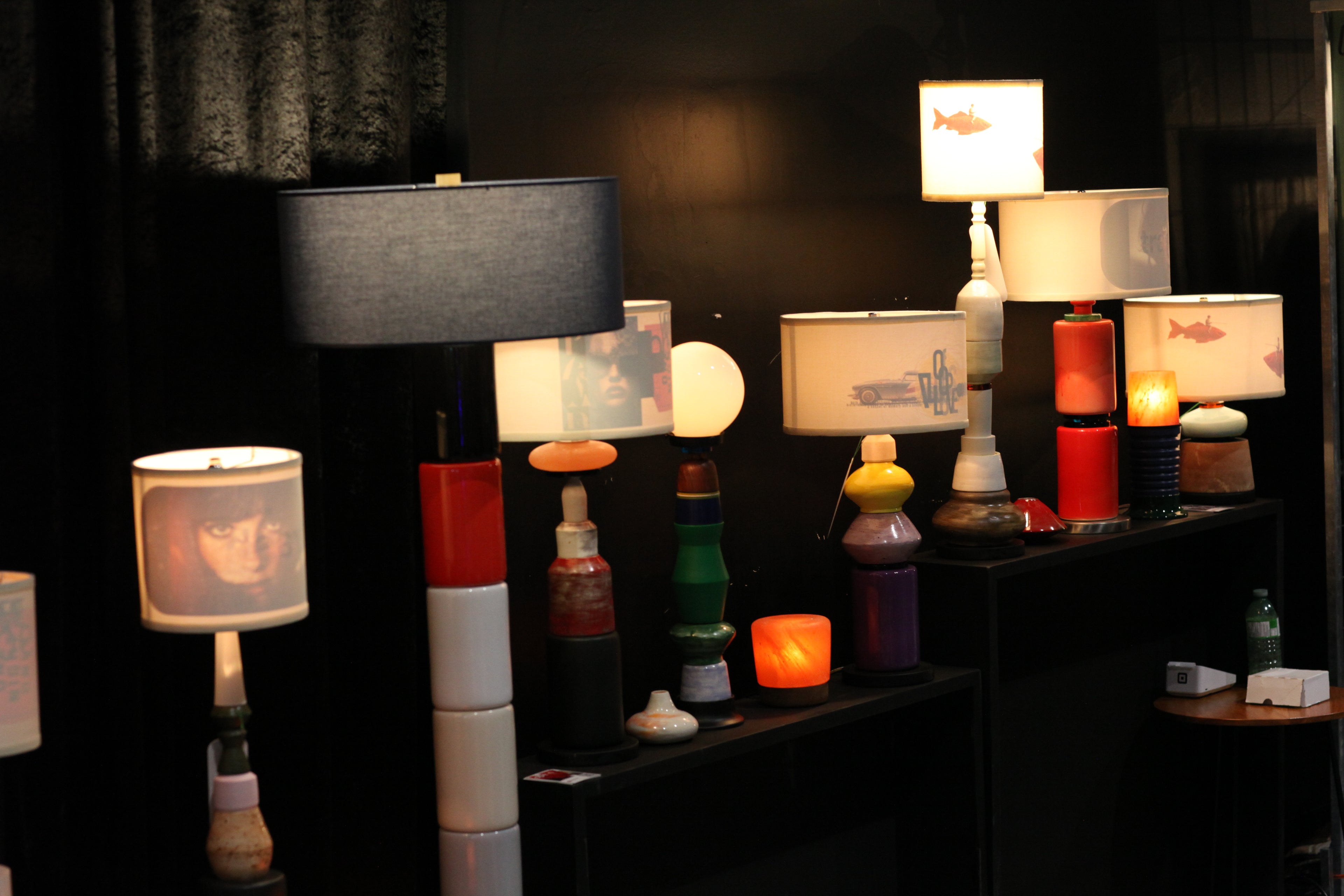 A collection of decorative table lamps displayed on black shelves against a dark background, including various lamp shades, colorful stacked bases, and lit lampshades.