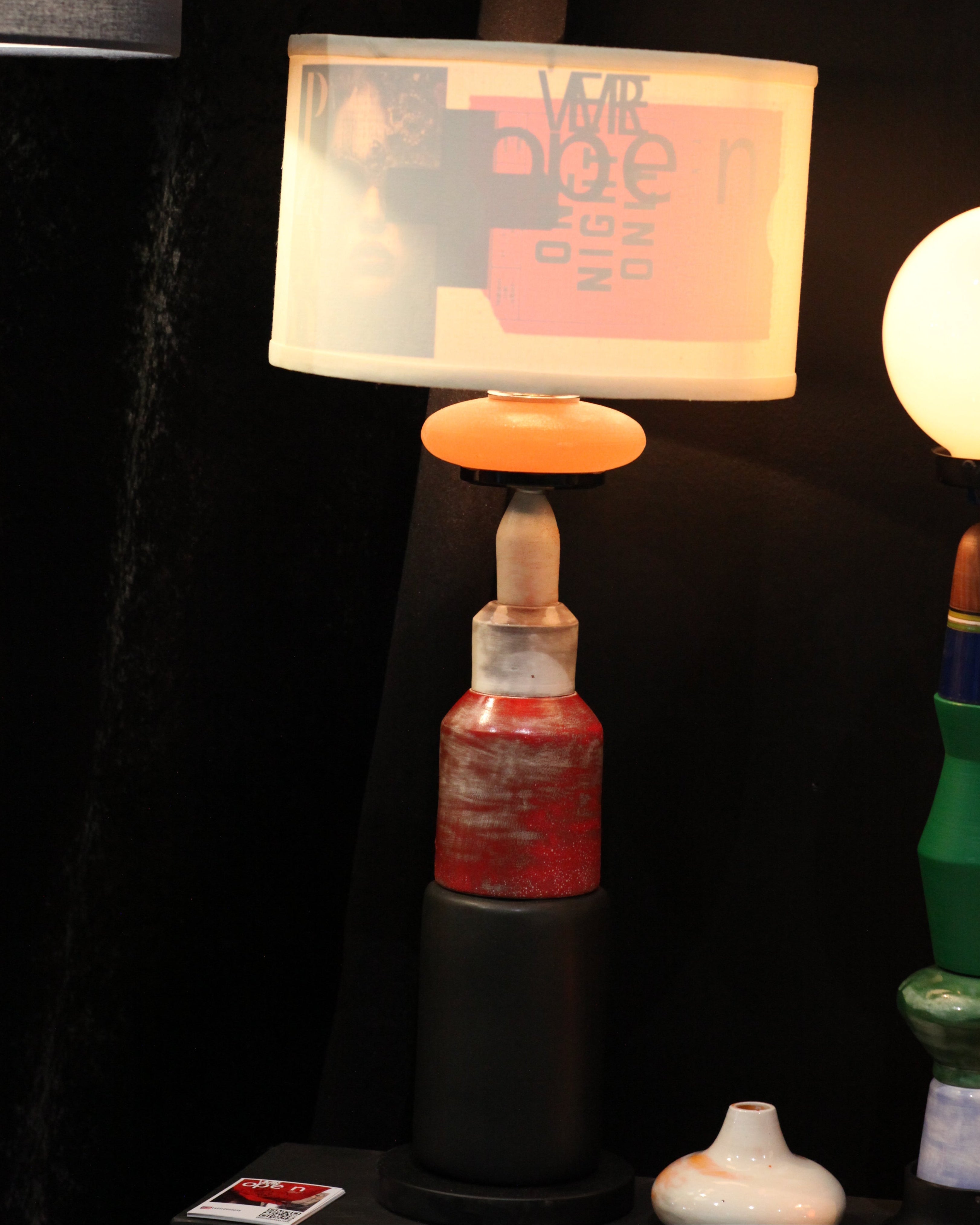 A close-up of a lamp with a translucent white shade featuring a pink graphic print, a glowing warm white bulb, and a decorative neck made from repurposed bottles in green, red, and black.