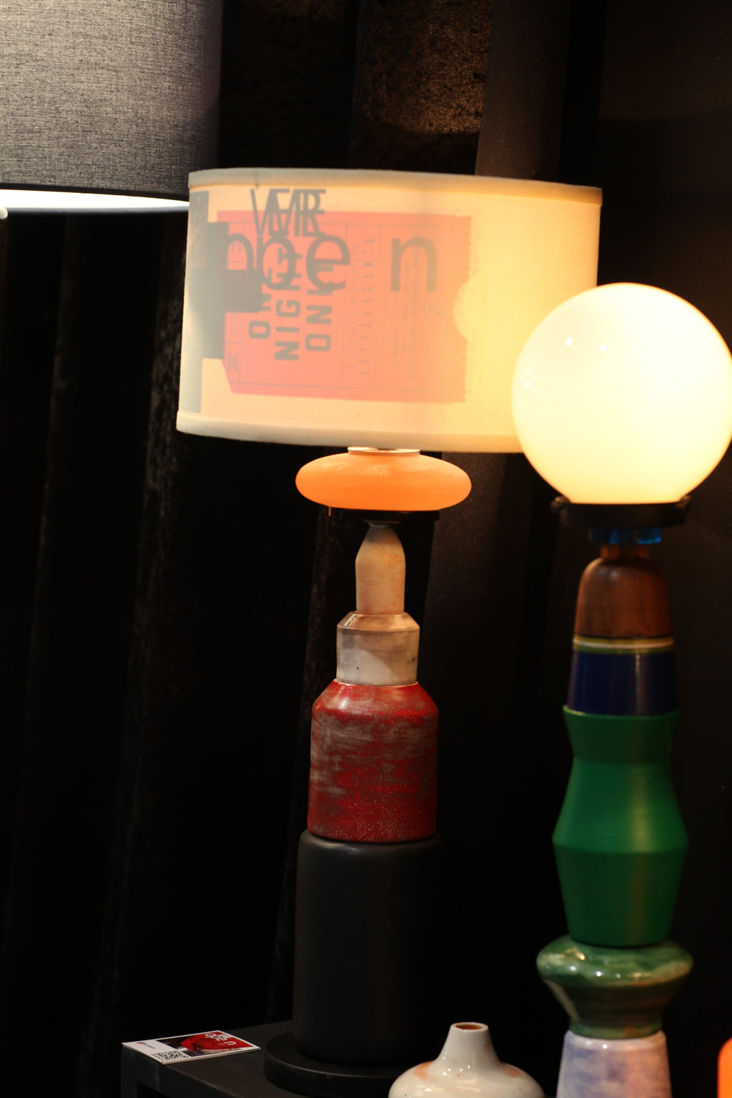 A close-up of a hanging light fixture with a glowing warm white globe bulb, a translucent white cylindrical shade printed with a pink graphic, and colorful metal hardware in green, red, and black against a dark background.