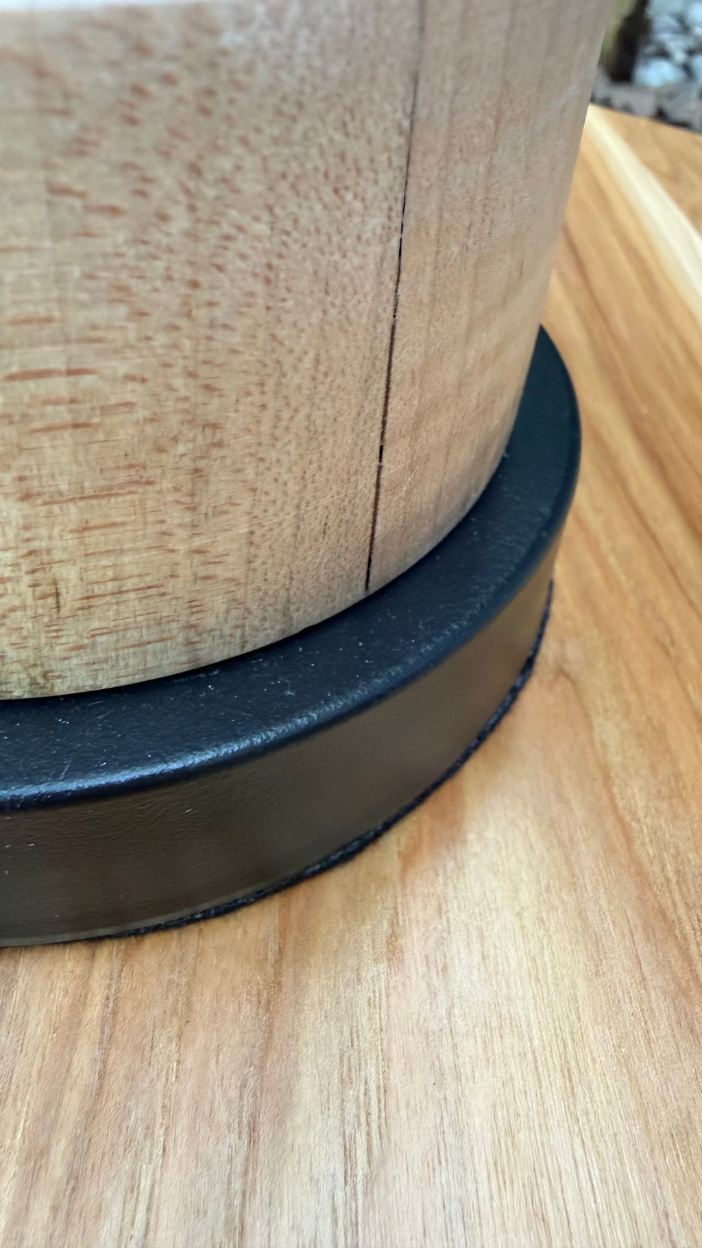 Close-up of the base of a cylindrical wooden item, showing a light natural wood grain surface and a wide black circular ring or base, photographed on a wood-grain surface.