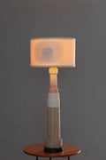 A modern table lamp with a cylindrical wooden base and a rectangular fabric shade glowing with a warm light, displayed on a small wooden side table against a dark gray background.