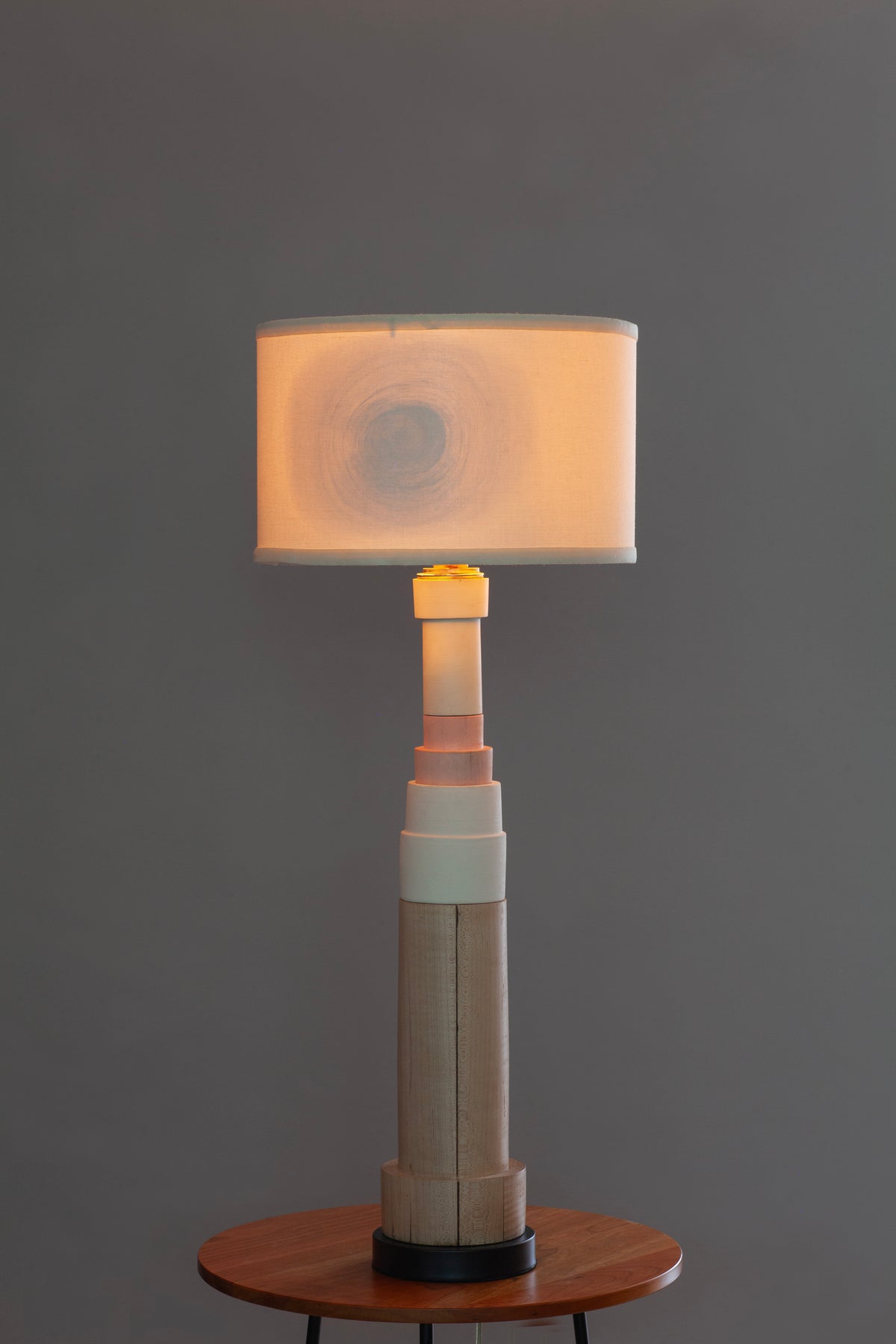 A modern table lamp with a cylindrical wooden base and a rectangular fabric shade glowing with a warm light, displayed on a small wooden side table against a dark gray background.