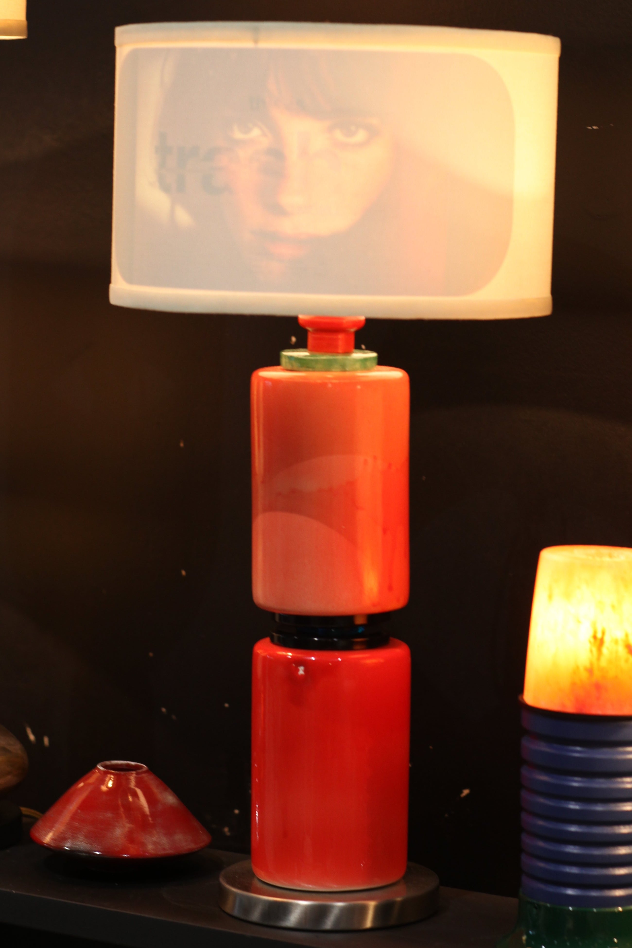 A close-up of a decorative table lamp with a translucent frosted square shade, red cylindrical body sections, and a dark blue ribbed base, photographed against a dark background.