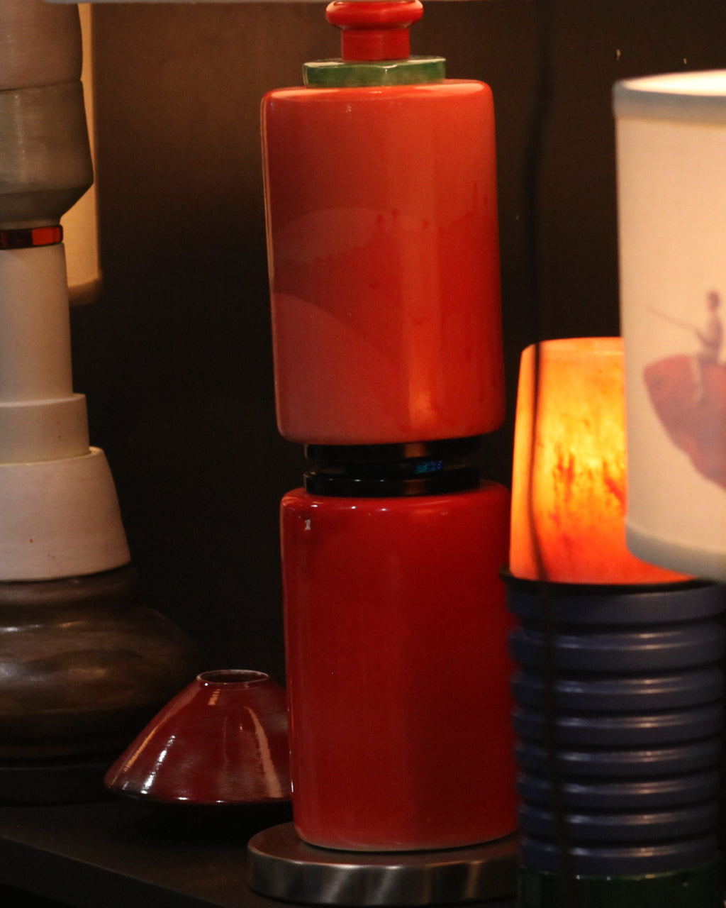 A collection of vintage and modern decorative lamps displayed on a dark shelf, featuring colorful ceramic bases, white drum shades, and one lamp with a fish graphic on the shade.