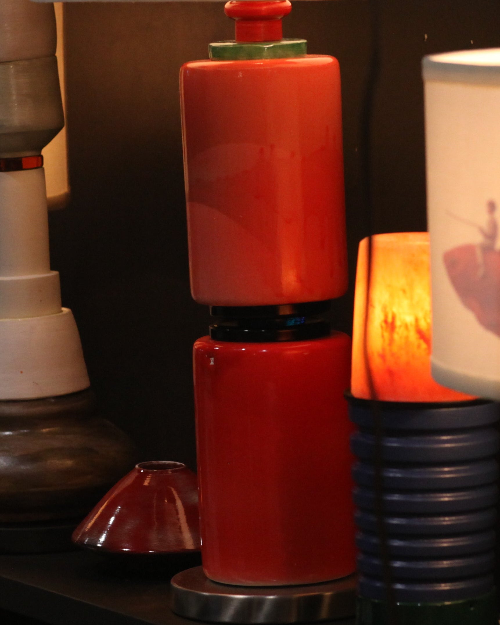 A collection of vintage and modern decorative lamps displayed on a dark shelf, featuring colorful ceramic bases, white drum shades, and one lamp with a fish graphic on the shade.