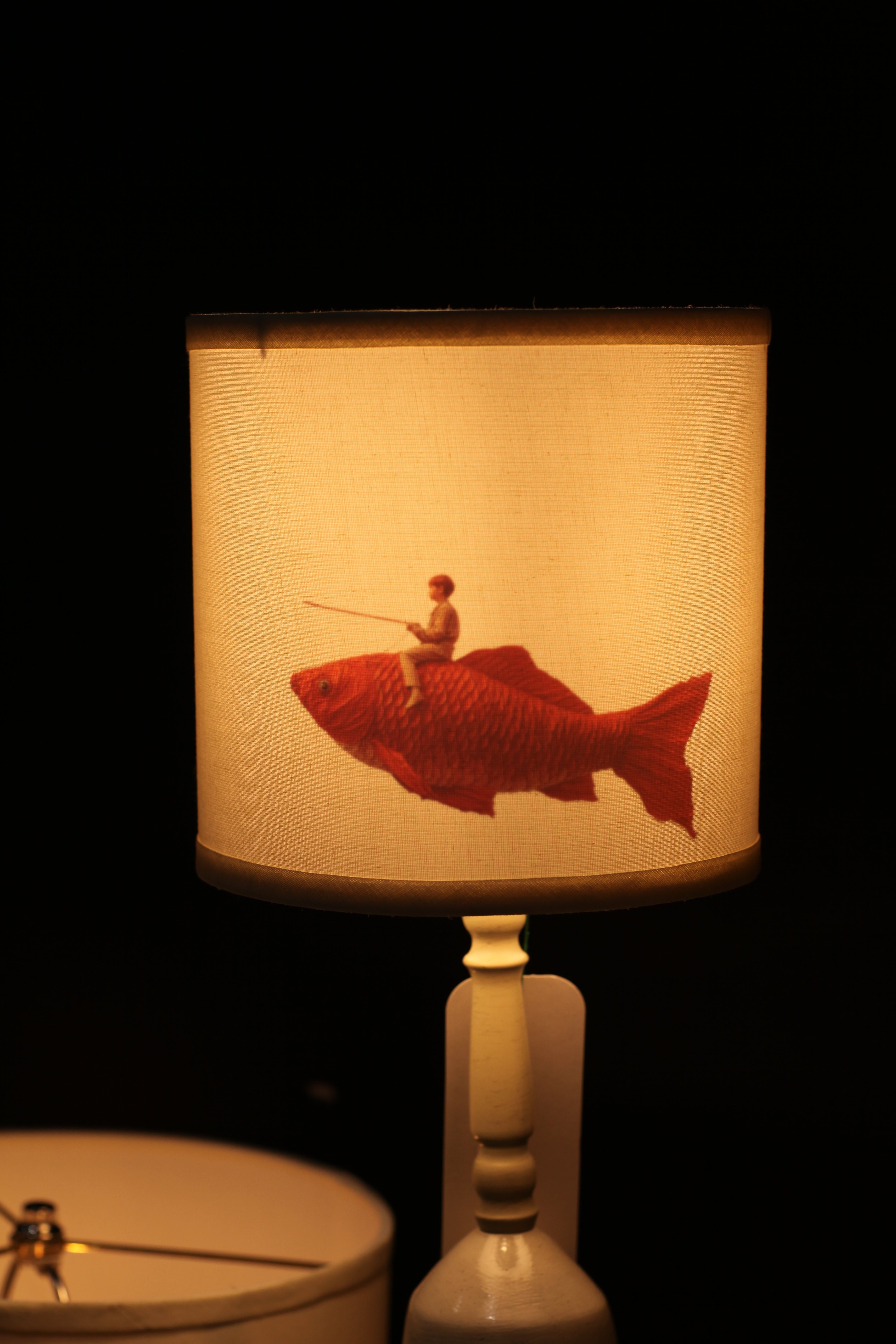 A cylindrical fabric lampshade with a printed illustration of a red fish holding a fishing rod, shown illuminated against a dark background.