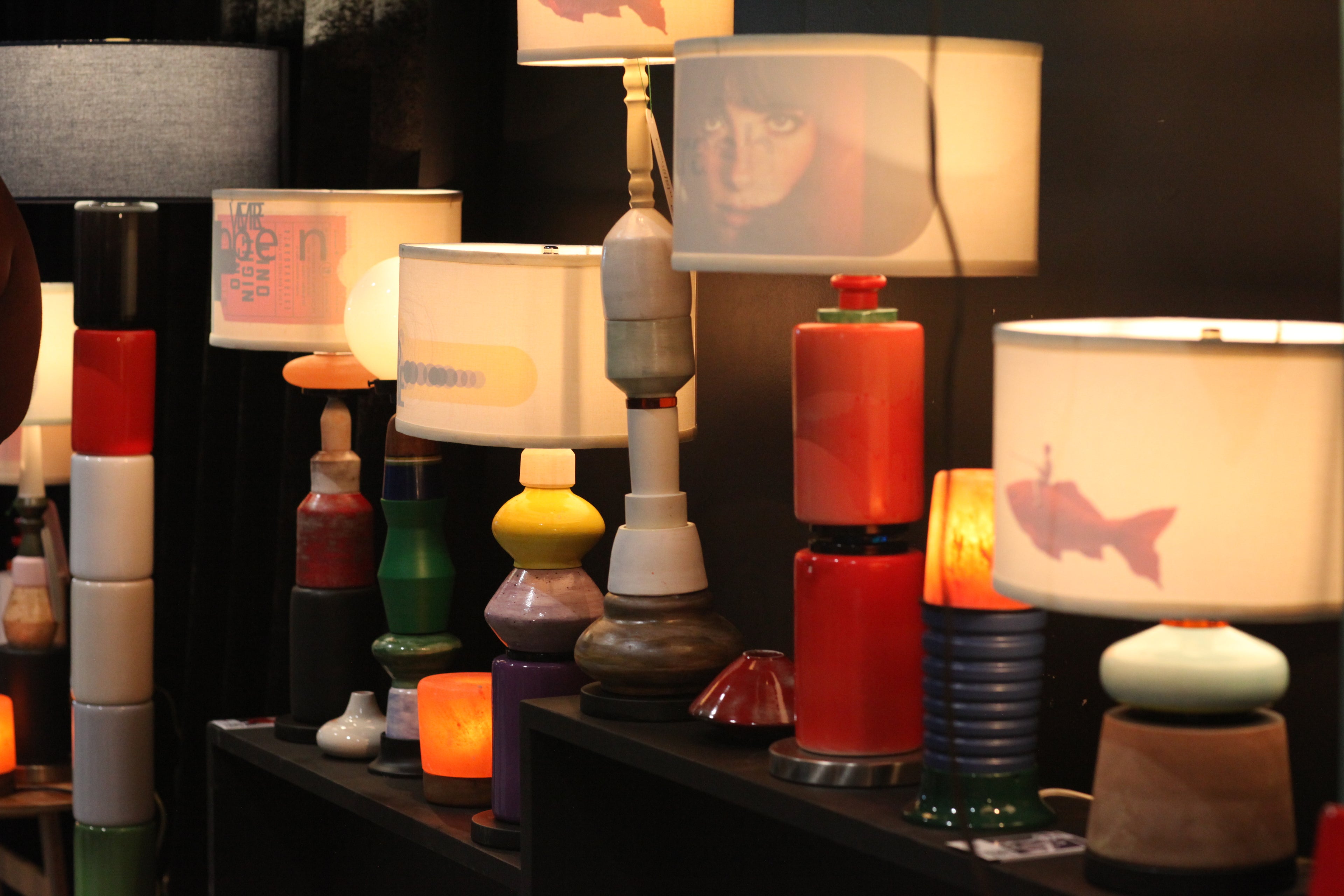 A collection of unique, colorful sculptural table lamps displayed on a dark shelf in a dimly lit room. The lamps feature various modern designs, including stacked cylindrical bases in red, white, and black, and white drum shades with printed graphics.