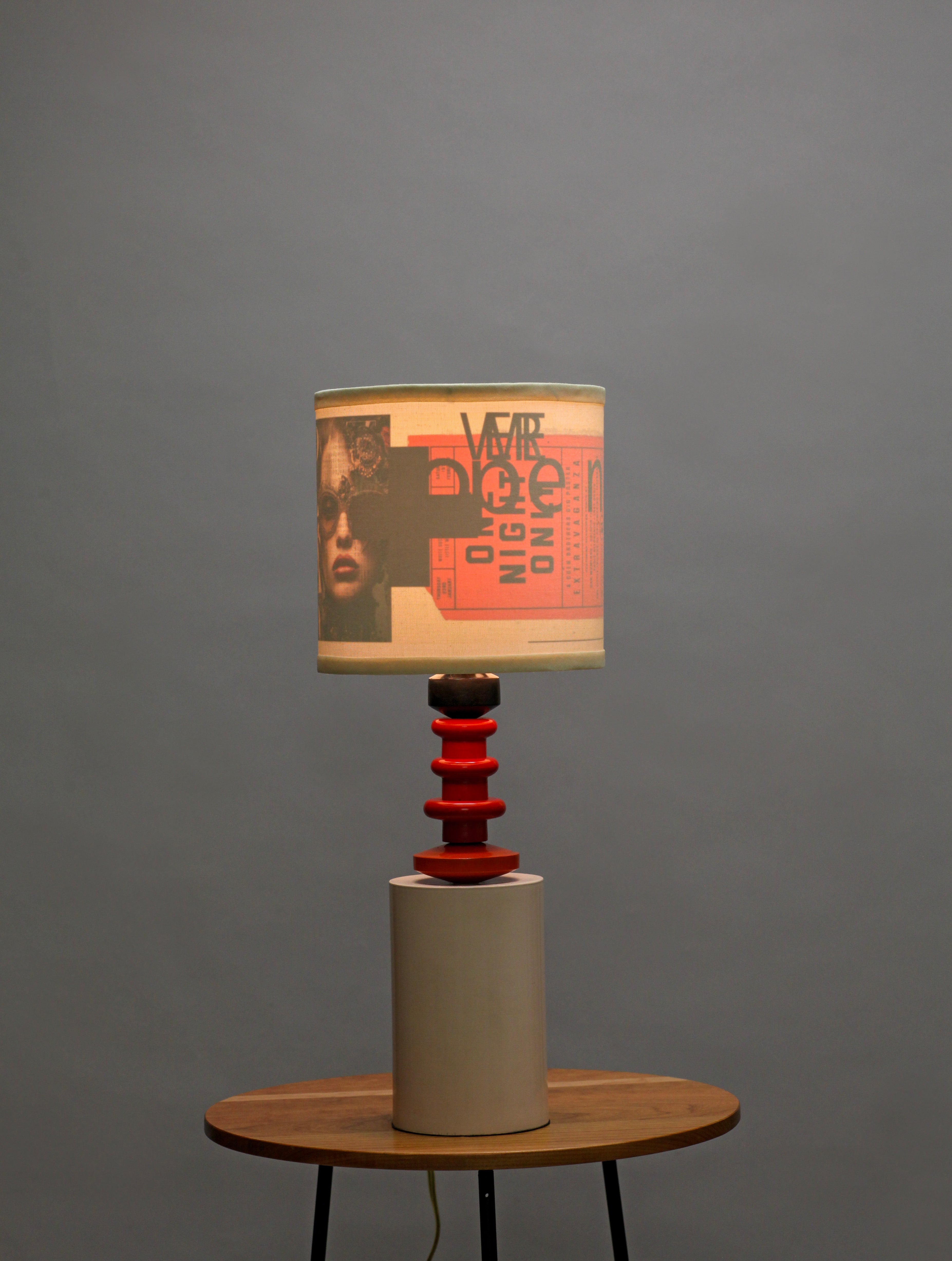 A table lamp with a beige drum shade featuring a graphic print of a face and the text 'VOTE DOCTOR NIGHT ONE', supported by a red spiral stem and a beige cylindrical base, set on a wooden table against a gray background.