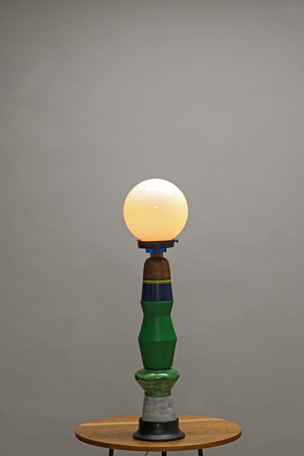 A vintage-style table lamp with a glowing warm white globe bulb, a sculptural green and blue ceramic base, and a small round wooden table against a plain gray studio background.