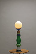 A vintage-style table lamp with a glowing warm white globe bulb, a sculptural green and blue ceramic base, and a small round wooden table against a plain gray studio background.
