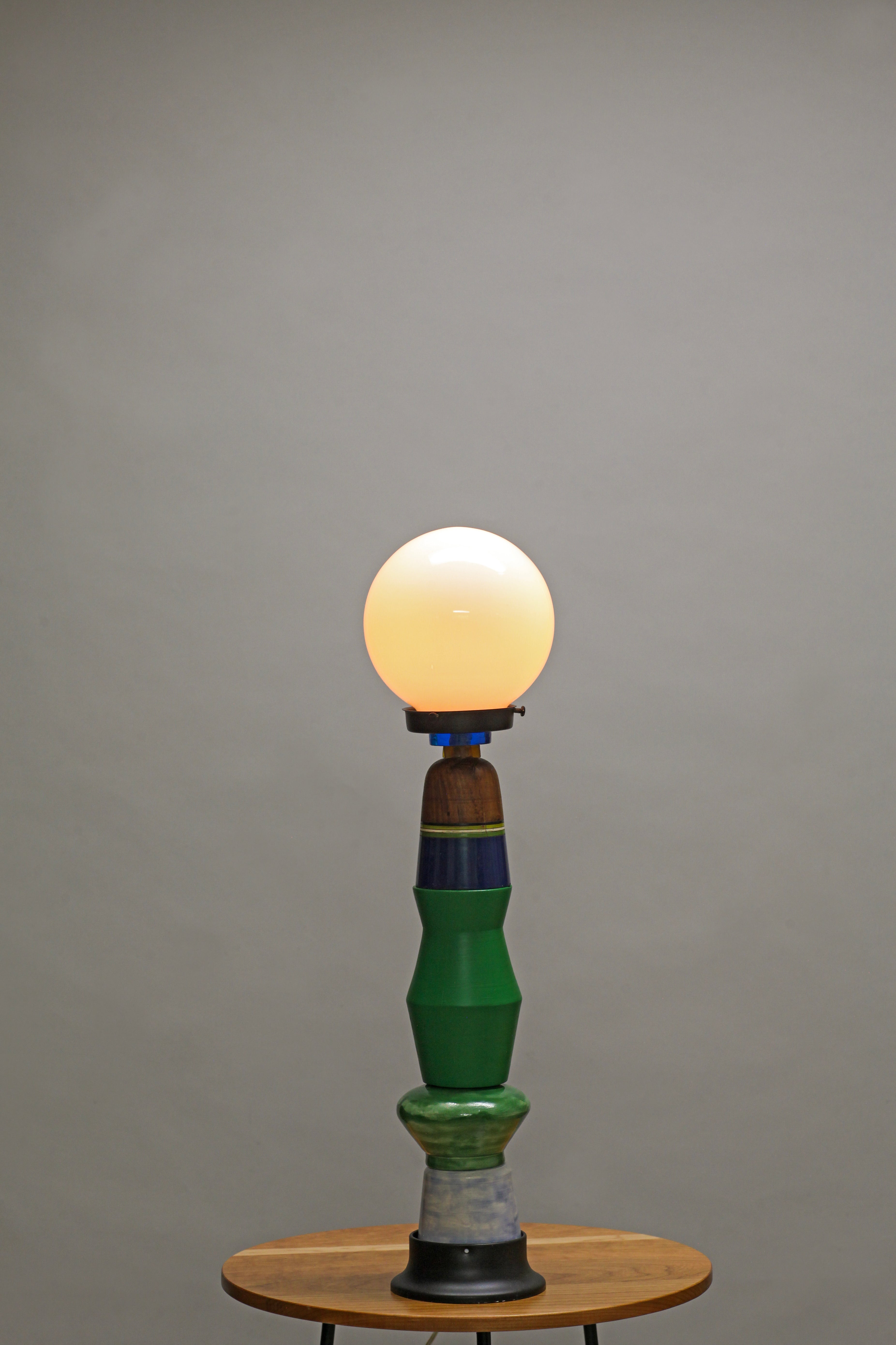 A vintage-style table lamp with a glowing warm white globe bulb, a sculptural green and blue ceramic base, and a small round wooden table against a plain gray studio background.