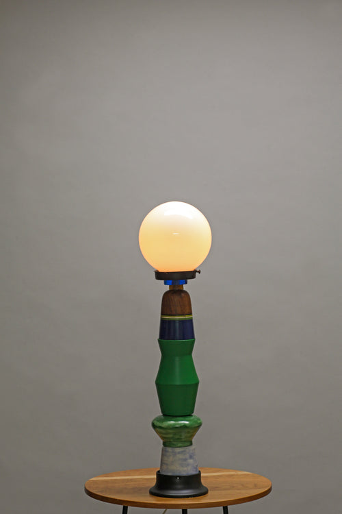 A vintage-style table lamp with a glowing warm white globe bulb, a sculptural green and blue ceramic base, and a small round wooden table against a plain gray studio background.